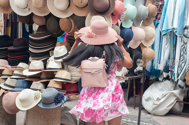 A young girl is seen from behind as she tries on a stylish pink hat amidst a vibrant display of various hats at an outdoor market stall.